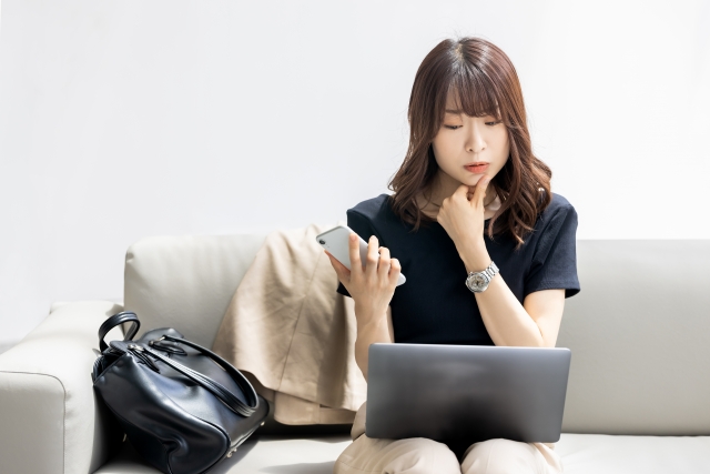 スマホを手にしながらパソコンを見、考える女性